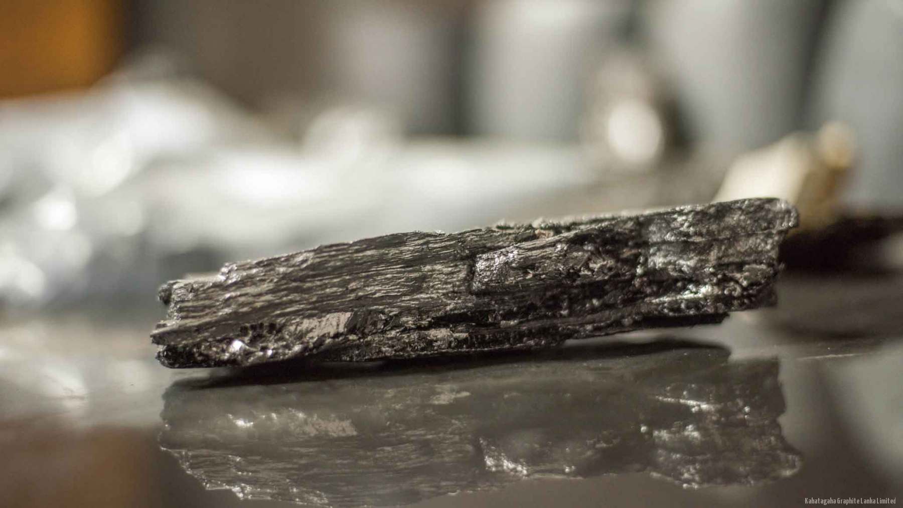 A single specimen of crystalline vein graphite resting on a reflective surface.